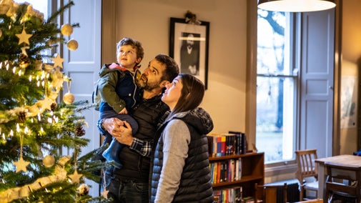Family of three with a young boy being lifted up to look at a Christmas tree inside a house.